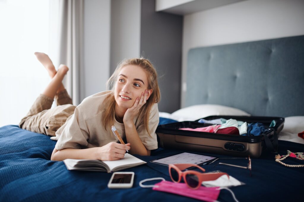 Young woman writing notes and packing clothes into suitcase indoors at home, planning holiday.